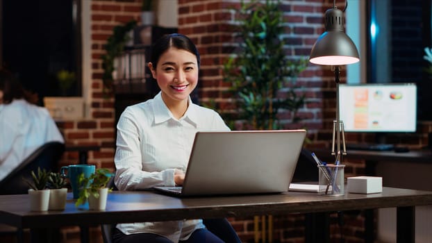 Portrait of smiling asian businesswoman doing computer tasks for team project in office. Cheerful employee working late at night, typing on laptop keyboard next to coworker, camera B