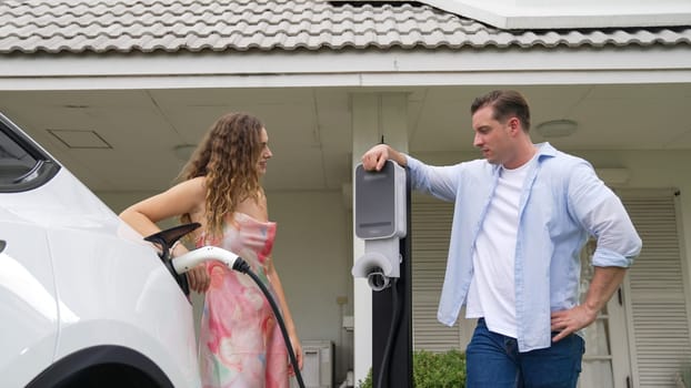 Happy and lovely couple with eco-friendly conscious recharging electric vehicle from EV charging station. EV car technology utilized as alternative transportation for future sustainability. Synchronos