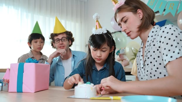 Caucasian mother cutting cake while family congratulate in girl's birthday. Diverse family celebrate daughter important day while waiting for eating dessert and food while smiling together. Pedagogy.