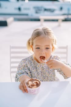 Little girl eats chocolate ice cream with a spoon from a cup while sitting at a table. High quality photo