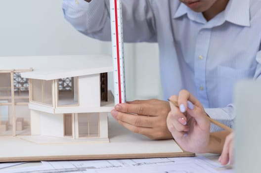 Professional male asian architect using ruler to measure house model length while young beautiful caucasian colleague using laptop to analyzed data on meeting table with house model. Immaculate.