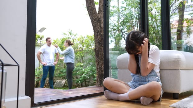 Stressed and unhappy young girl huddle in corner, cover her ears blocking sound of her parent arguing in background. Domestic violence at home and traumatic childhood develop to depression. Synchronos