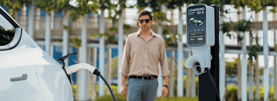 Young man recharge electric car's battery from charging station in outdoor green city park. Rechargeable EV car for sustainable environmental friendly urban travel. Panorama Expedient