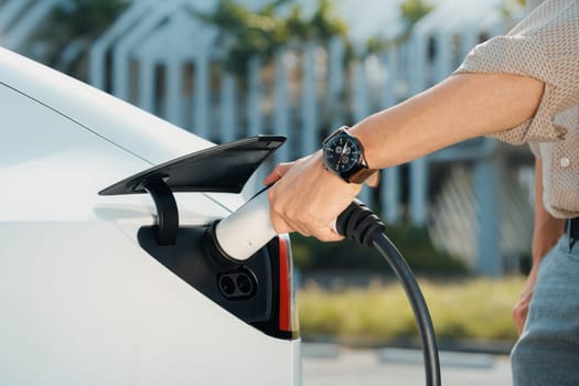 Young man put EV charger to recharge electric car's battery from charging station in city commercial parking lot. Rechargeable EV car for sustainable environmental friendly urban travel. Expedient