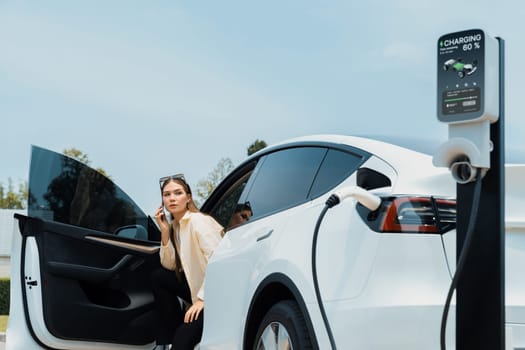 Young woman use smartphone to pay for electricity at public EV car charging station green city park. Modern environmental and sustainable urban lifestyle with EV vehicle. Expedient