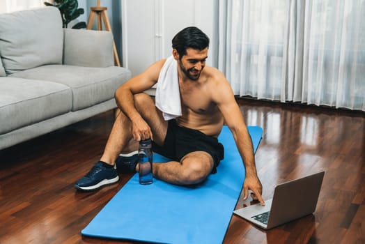 Athletic and sporty man resting on fitness mat during online body workout exercise session for fit physique and healthy sport lifestyle at home. Gaiety home exercise workout training concept.