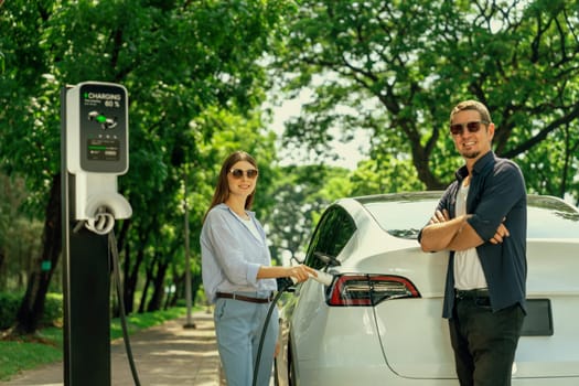 Lovely young couple wearing sun glasses recharging battery for electric car during road trip travel EV car in natural forest or national park. Eco friendly travel during vacation and holiday. Exalt