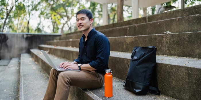 Young businessman working online with laptop while sitting on bench outside of office building.