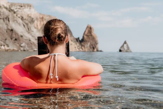 Woman freelancer works on laptop swimming in sea on pink inflatable ring. Pretty lady typing on computer while floating in the sea on inflatable donut at sunset. Freelance, remote work on vacation