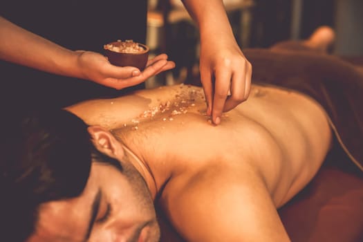 Man customer having exfoliation treatment in luxury spa salon with warmth candle light ambient. Salt scrub beauty treatment in Health spa body scrub. Quiescent