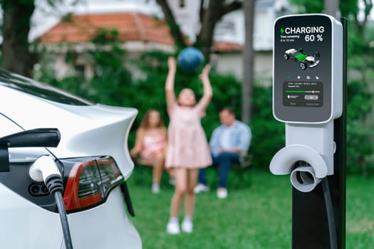 Focus electric vehicle recharge from home charging station on blur background of happy and playful family playing together. EV car using alternative and sustainable energy for better future.Synchronos