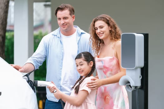 Happy little young girl learn about eco-friendly and energy sustainability as she help her family recharge electric vehicle from home EV charging station. EV car and modern family concept. Synchronos