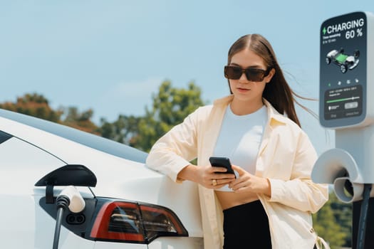 Young woman use smartphone to pay for electricity at public EV car charging station green city park. Modern environmental and sustainable urban lifestyle with EV vehicle. Expedient