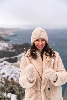 Outdoor winter portrait of happy smiling woman, light faux fur coat holding heart sparkler, posing against sea and snow background.