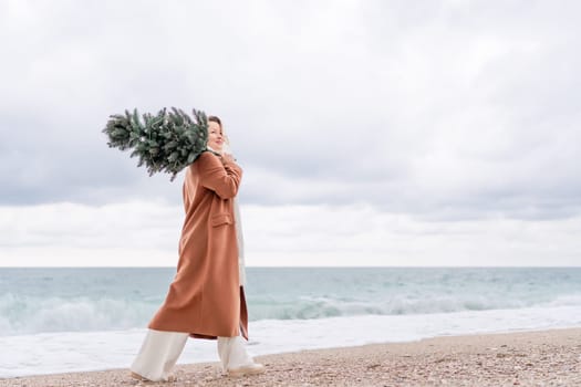 Blond woman Christmas sea. Christmas portrait of a happy woman walking along the beach and holding a Christmas tree on her shoulder. She is wearing a brown coat and a white suit