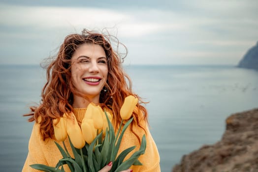 Portrait of a happy woman with hair flying in the wind against the backdrop of mountains and sea. Holding a bouquet of yellow tulips in her hands, wearing a yellow sweater.