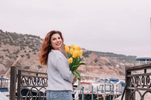 Woman holds yellow tulips in harbor with boats docked in the background., overcast day, yellow sweater, mountains.