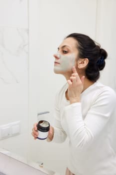 Caucasian woman looking in mirror and applying face mask white in bathroom