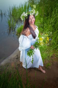 Adult mature brunette woman in a white dress, sundress and a wreath of flowers in summer by the water of river or lake in evening at sunset. Celebration of the Slavic pagan holiday of Ivan Kupala