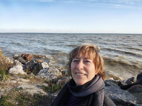 Happy cheerful middle aged woman in scarf taking selfie on nature outdoors and sun with water and waves of sea on the background