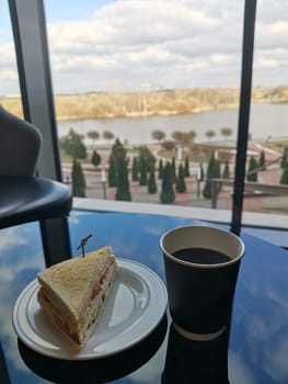 Paper cup of coffee and sandwich in a cafeteria on a glass table with a reflection of the sky.