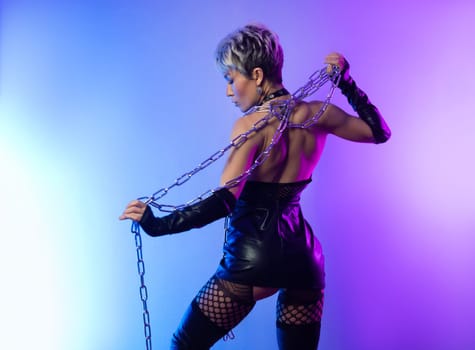 sexy girl in the erotic image of a bdsm mistress in leather belts with a metal chain shackles poses against a background of copy paste in a beautiful neon background of copy paste