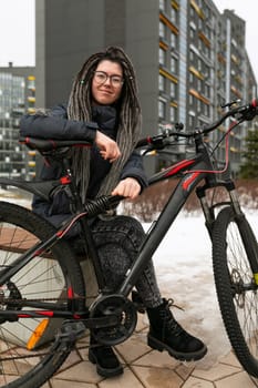 Photo on the street, a young active woman with a dreadlocked hairstyle, dressed in a warm jacket, rides a bicycle.