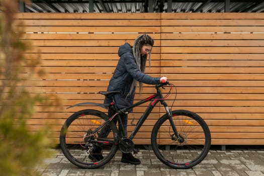 Cute young woman with dreadlocks hairstyle dressed in a winter jacket rides a bicycle.