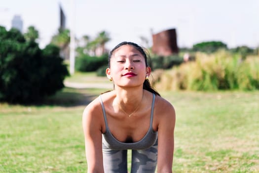 front view of a young asian woman doing yoga stretching exercises in the grass of the park, active and healthy lifestyle concept