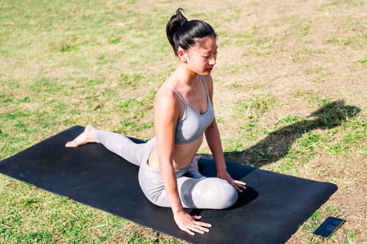 young asian woman doing stretching exercises with her yoga mat on the grass in the park using the app on her mobile phone, active and healthy lifestyle concept