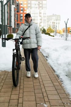 A young Caucasian man with a gray jacket and black cap is waiting for a friend with a bicycle under the porch.