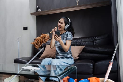 Funny woman Asian teenage cleaning house, doing housework, cleaning the floor with duster and listening to music in wireless headphones.