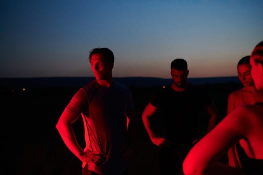 In the late-night hours, a diverse group of exhausted athletes find solace under a red glow, reflecting on their day-long marathon journey and celebrating camaraderie amidst fatigue.