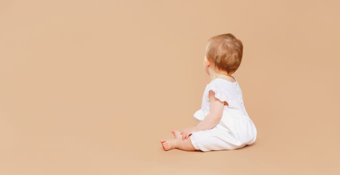 Happy cute baby sitting on the floor on brown studio background, back view, blank copy space for advertising text