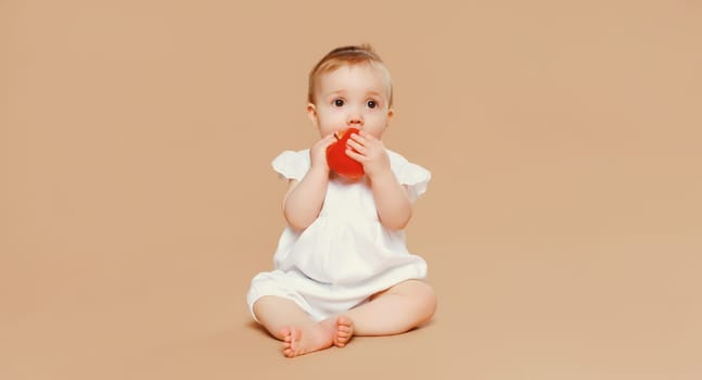 Portrait of cute baby holding red apple fruit sitting on the floor on brown studio background