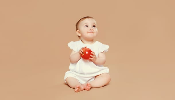 Portrait of cute baby holding red apple fruit sitting on the floor on brown studio background