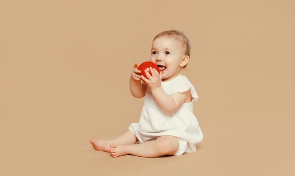 Portrait of cute baby holding red apple fruit sitting on the floor on brown studio background