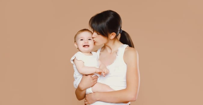 Happy young mother holding and kissing baby on brown studio background