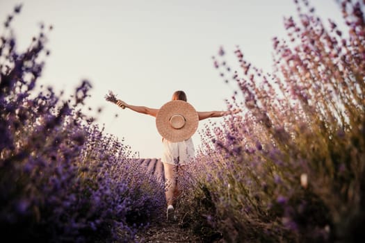 A woman is walking through a field of purple flowers with a straw hat on.