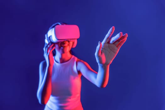 Smart female standing with surrounded by cyberpunk neon light wear VR headset connecting metaverse, futuristic cyberspace community technology. Woman using hand touching virtual object. Hallucination.