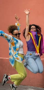 Two multiethnic women jumping in the air excited with rainbow flag in Madrid urban city street.