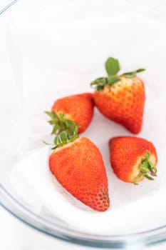 Bright red strawberries, interspersed with signs of mold, rest in a glass bowl lined with a paper towel on a white napkin, indicating improper storage techniques.