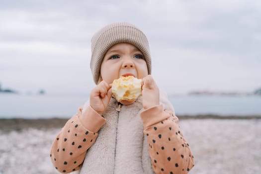 Little girl gnaws a big apple while holding it in her hands on the seashore. High quality photo