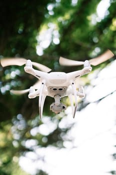 White four-engine drone hovers in the air in a green park. High quality photo