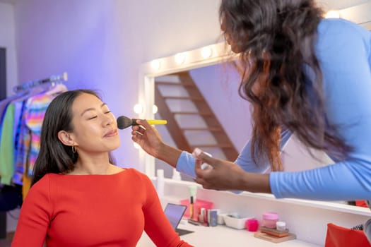 Woman with long hair is sharing a fun moment applying makeup to another woman's face in front of a mirror, Both enjoying the leisure of this artful gesture