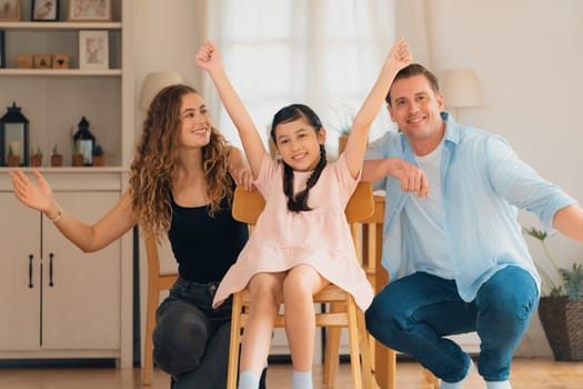 Happy family portrait with lovely little girl smiling and looking at camera, lovely and cheerful parent and their daughter sitting together in living room at home with warm daylight. Synchronos