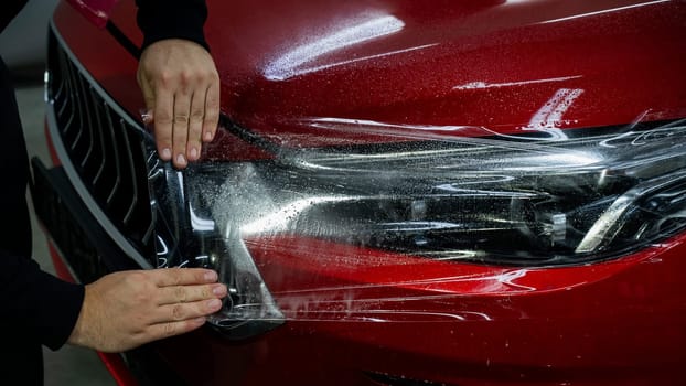 The master applies vinyl film to the headlight of a red car