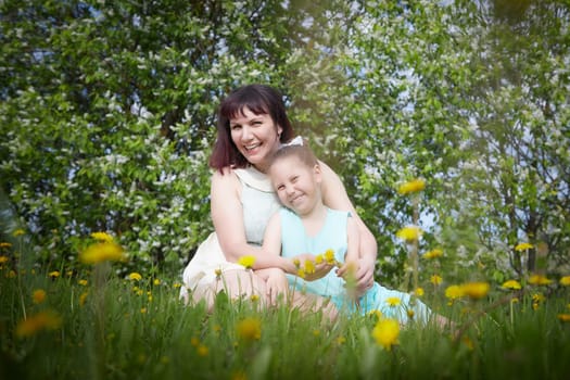 Happy mother and daughter enjoying rest, playing and fun on nature on a green lawn with dandelions and blooming apple tree on background. Woman and girl resting outdoors in summer and spring day