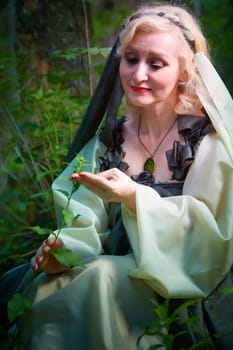 Adult mature woman 40-60 in a green long fairy dress in forest. Photo shoot in style of dryad and queen of nature. Fairy in beautiful green summer forest. Concept of caring for nature