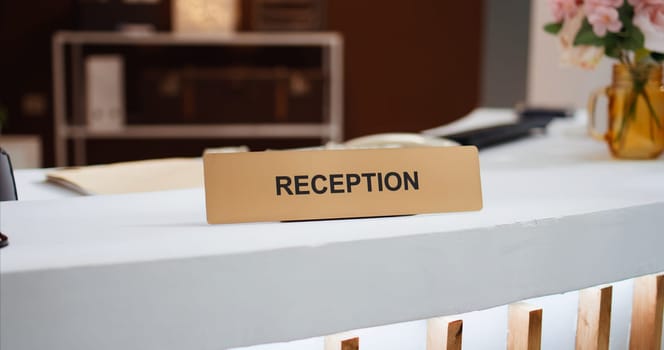 Reception sign placed on front desk in hotel lobby indicating place to book rooms and do check in process for accommodation. Empty resort hallway used to greet customers. Close up.